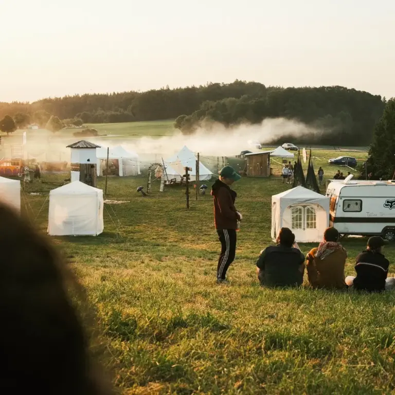 Festivalgäste auf der weitläufigen Campingwiese des DachüberKopf bei goldenem Abendlicht, Zelte und Wohnwagen im Hintergrund, Waldhorizont in der Ferne